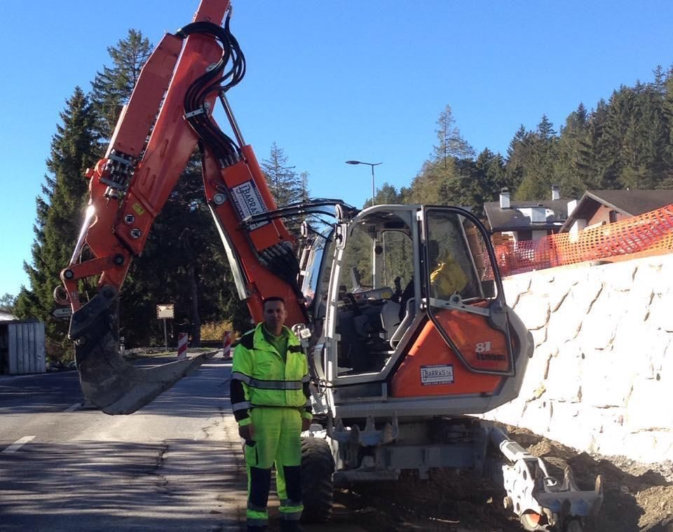 Une pelleteuse orange dans une rue, à côté d'un ouvrier en tenue de sécurité vert fluo et d'un mur de soutènement en pierre.