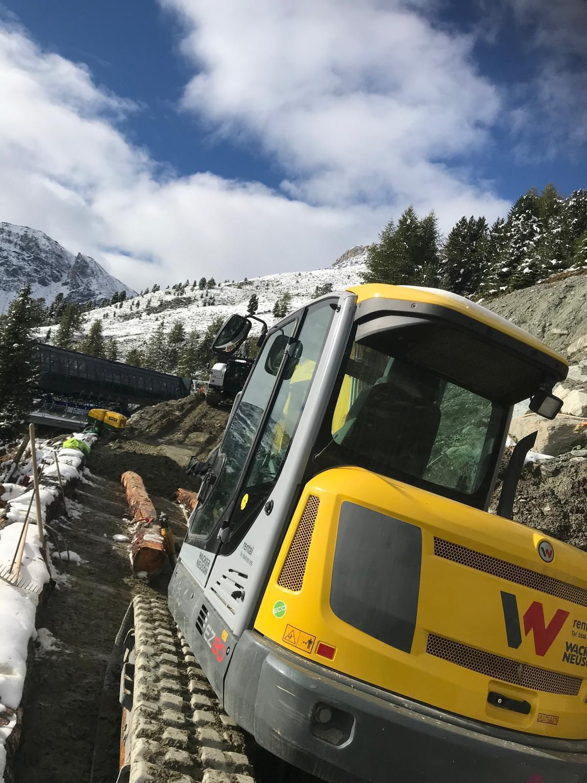 Une pelleteuse jaune sur un versant de montagne enneigé sous un ciel nuageux, avec une personne dans la cabine.
