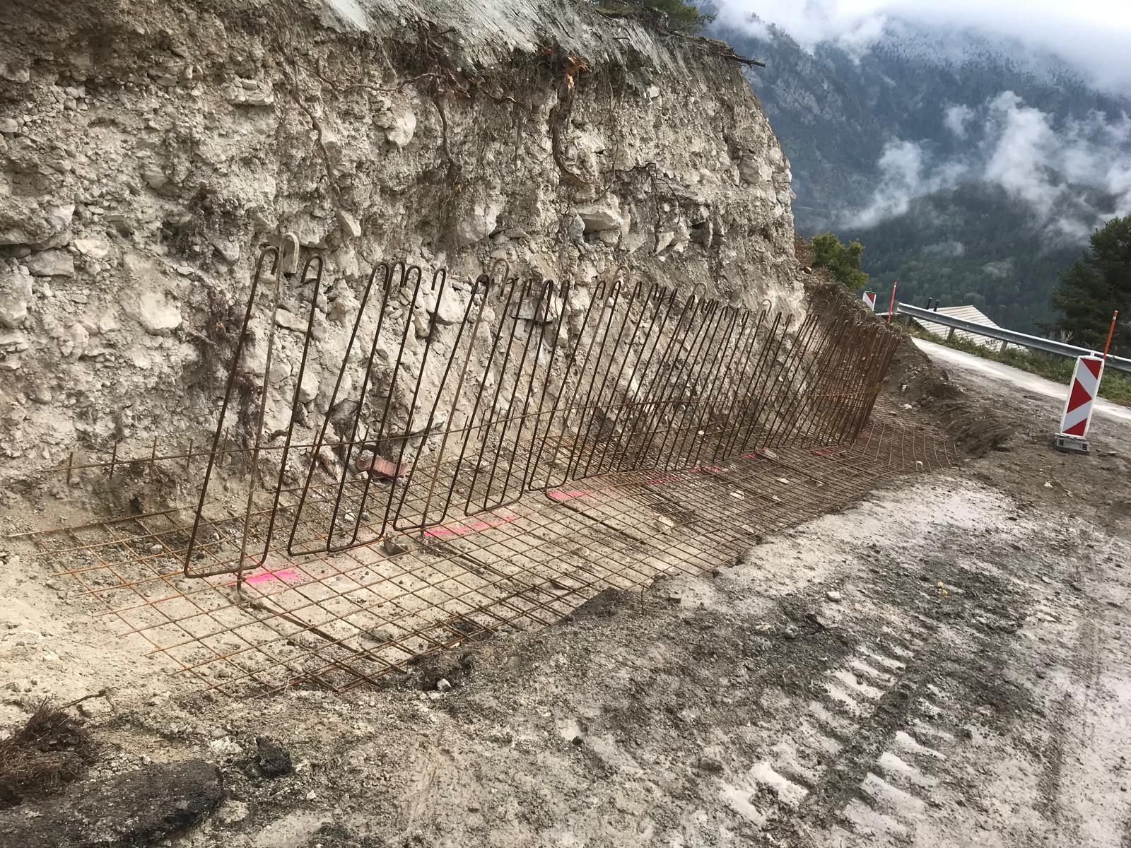 Route rocailleuse taillée avec des barres d'armature apparentes et un chemin de chantier boueux au bord de montagnes brumeuses