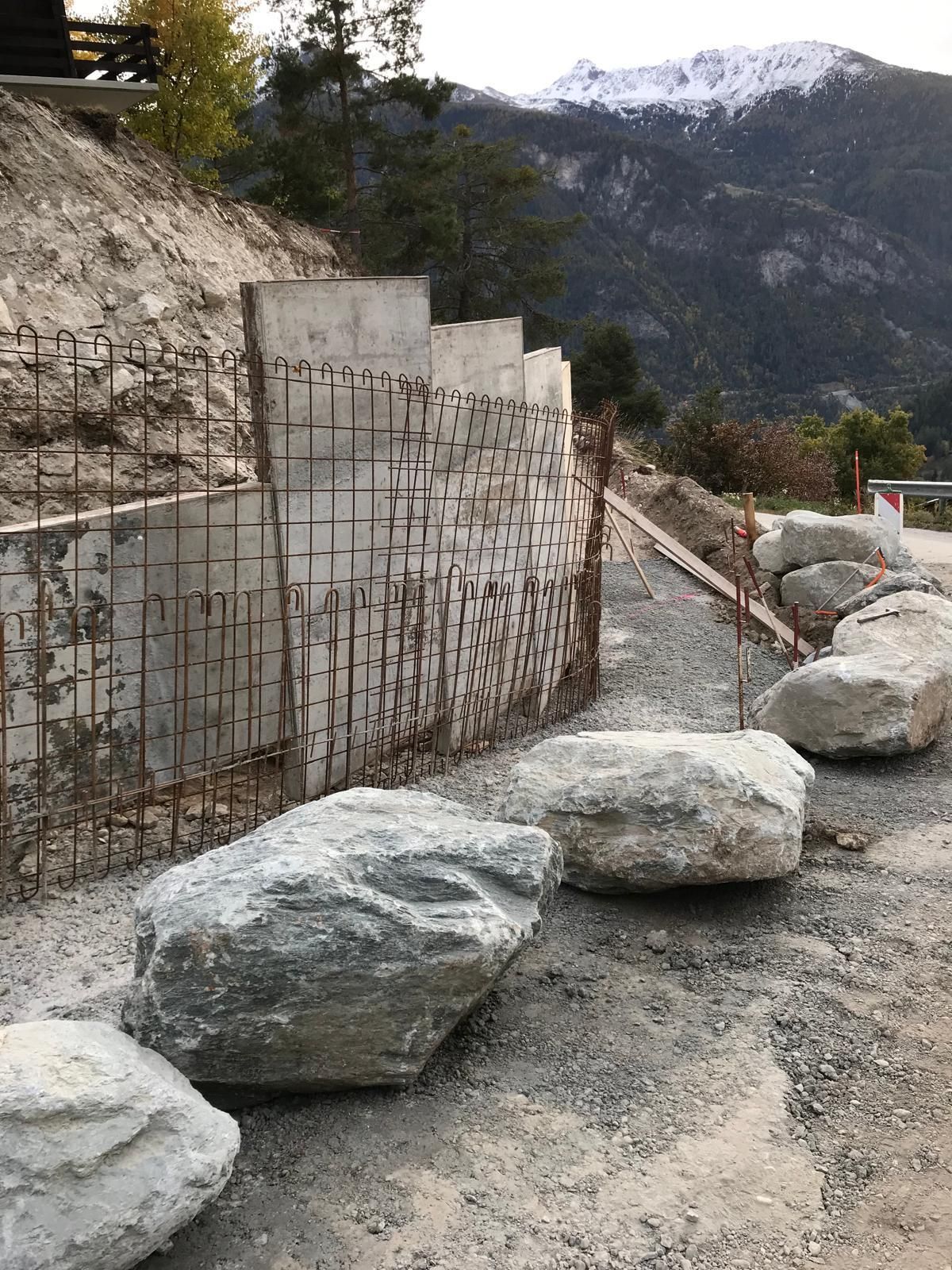 Chemin de gravier longeant un versant de montagne parsemé de gros rochers, de grillage et de sommets enneigés à l'horizon.