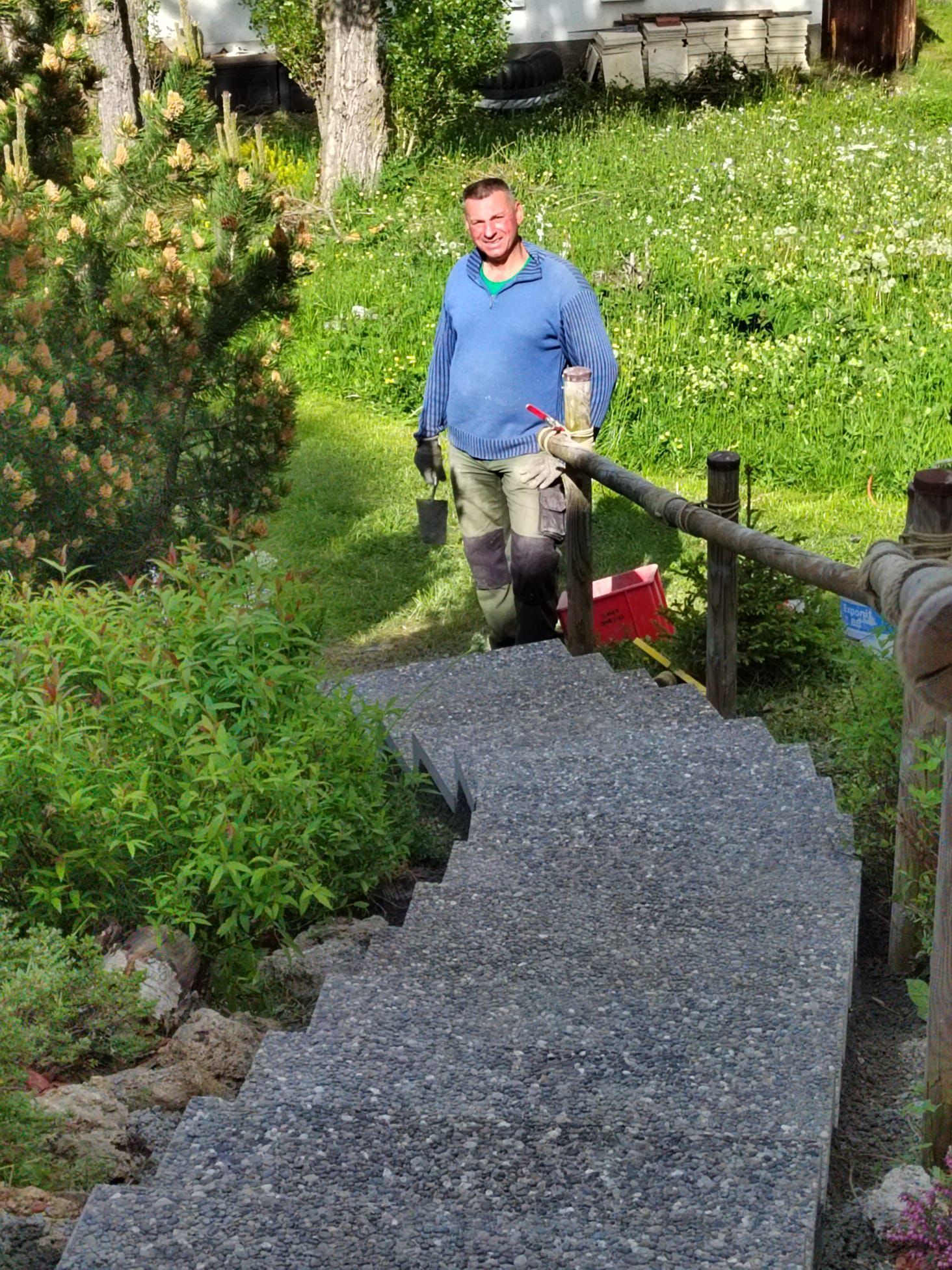 Une personne remonte un chemin de gravier dans un jardin, longeant une balustrade en bois, tenant un seau rouge.