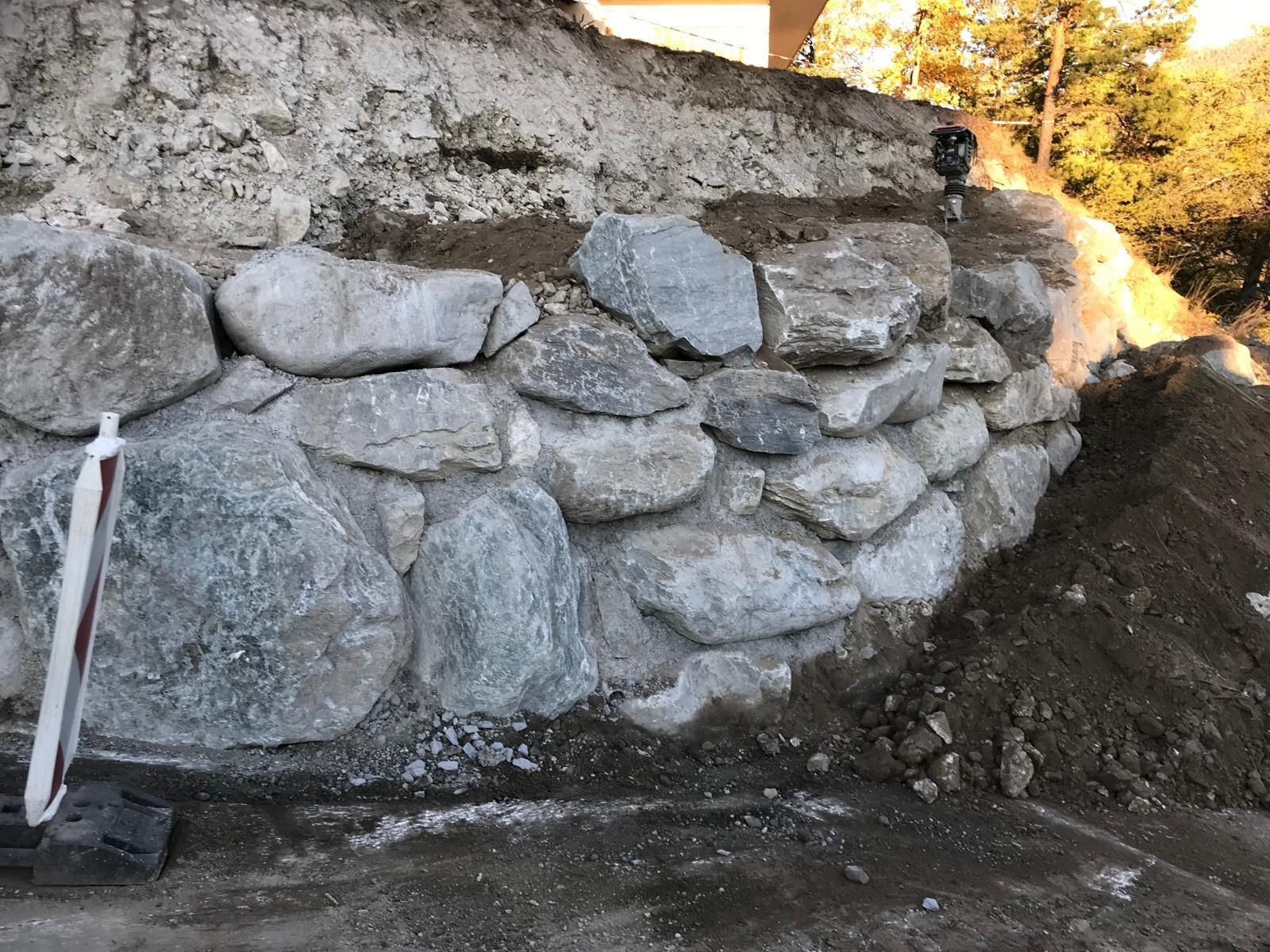 Mur de soutènement en pierre entouré de roches grises brutes et de terre, à côté d'une pelle sur la gauche