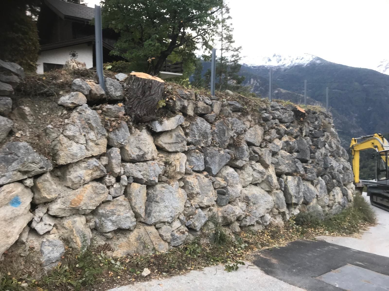 Mur de soutènement en pierre au bord d'une route de montagne, avec une excavatrice jaune en arrière-plan.