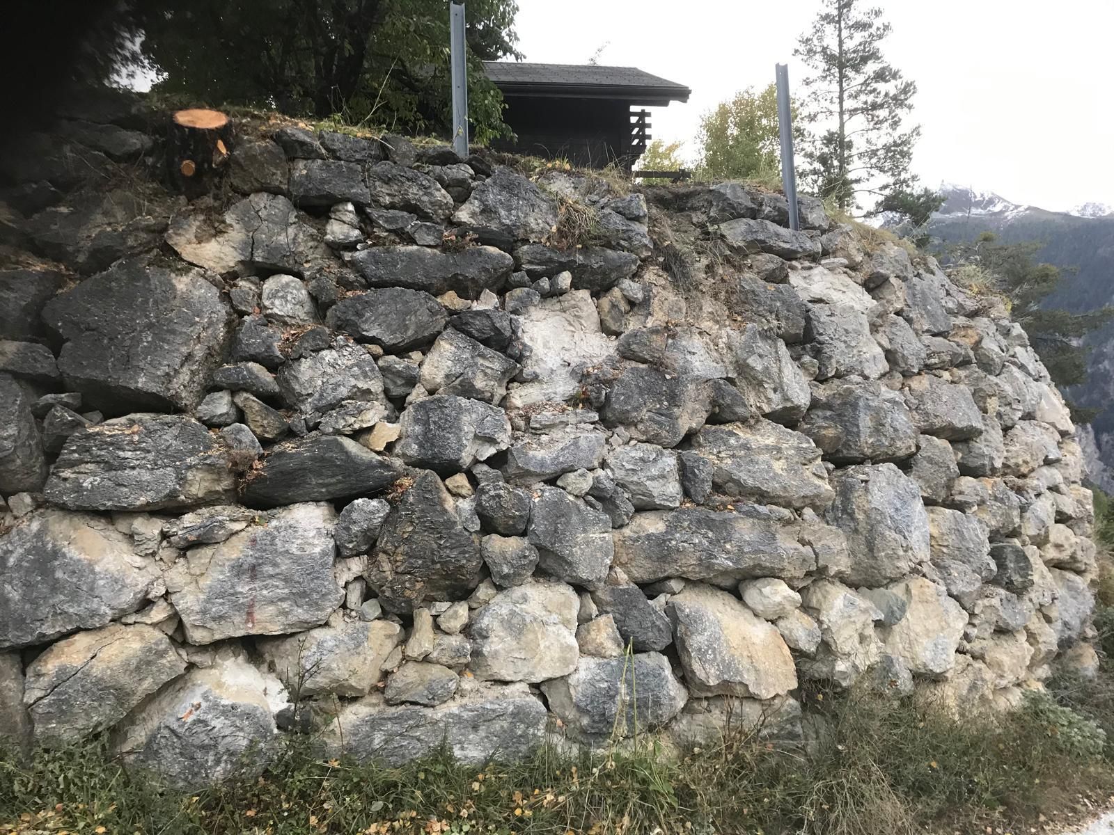 Mur de soutènement en pierre brute sur un flanc de colline, surmonté d'une petite structure couverte.