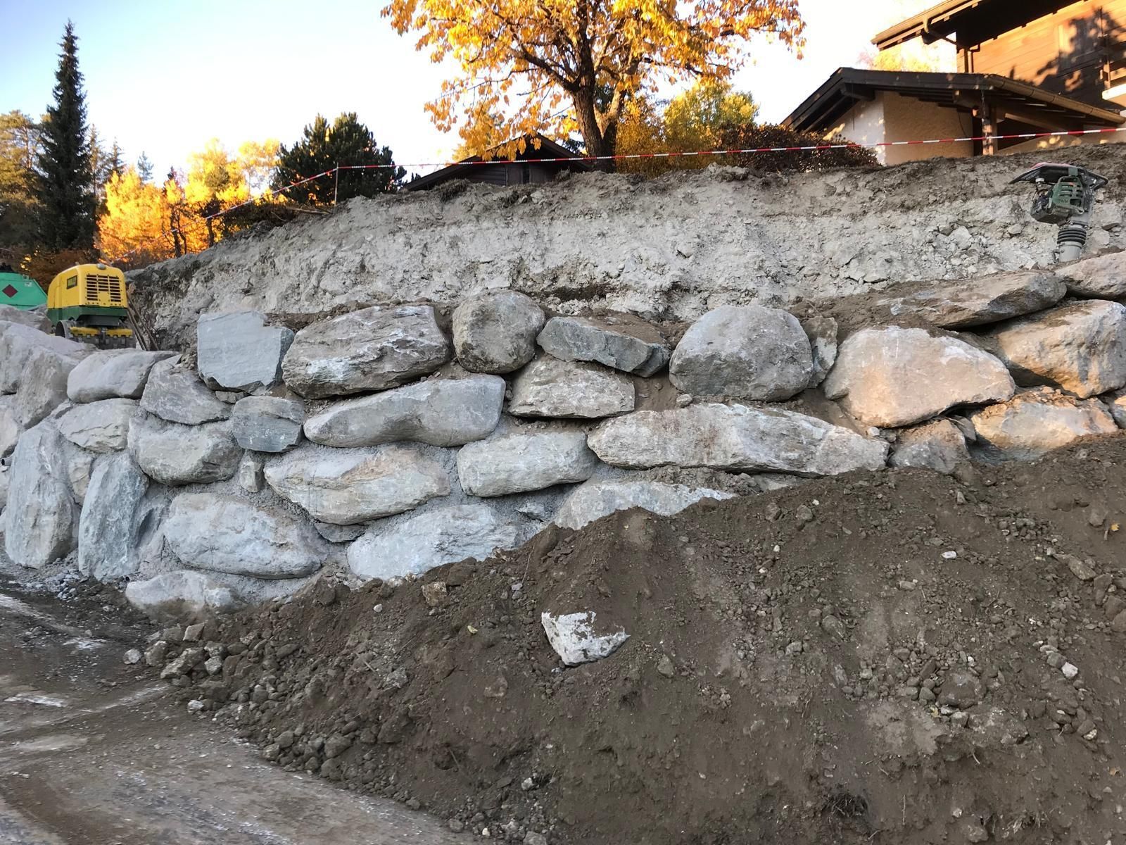 Mur de soutènement en pierre le long d'un talus de terre, avec des arbres d'automne et une maison en arrière-plan.