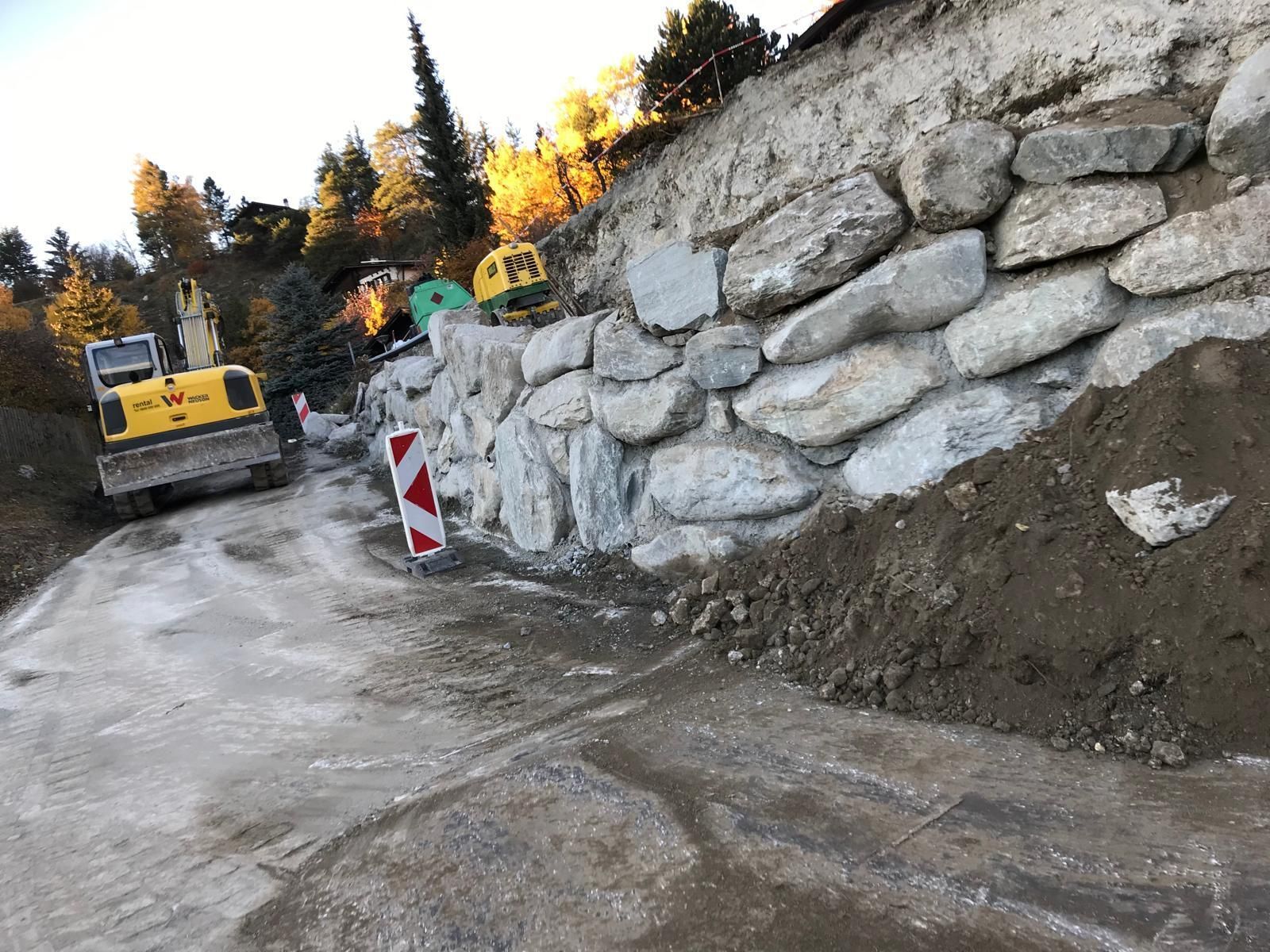 Chantier de construction routière avec une excavatrice jaune à côté d'un mur de soutènement en pierre et d'un remblai de terre.