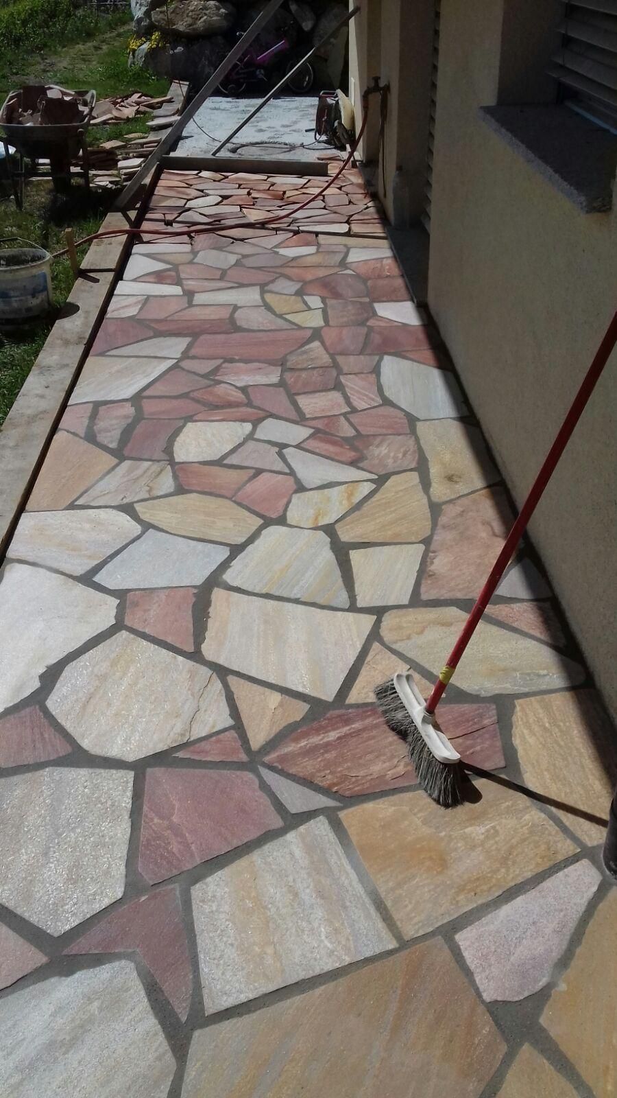 Allée carrelée longeant une maison, avec un balai à manche rouge appuyé contre le mur.