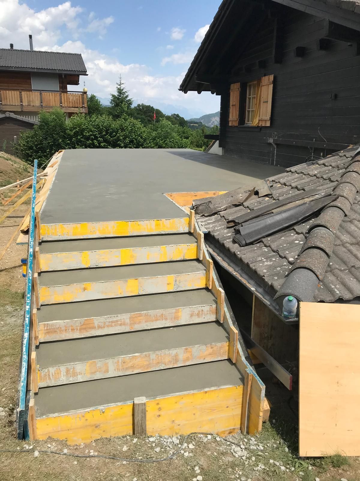 Rampe et escaliers en béton en construction à côté d'une maison en bois sombre au toit gris.