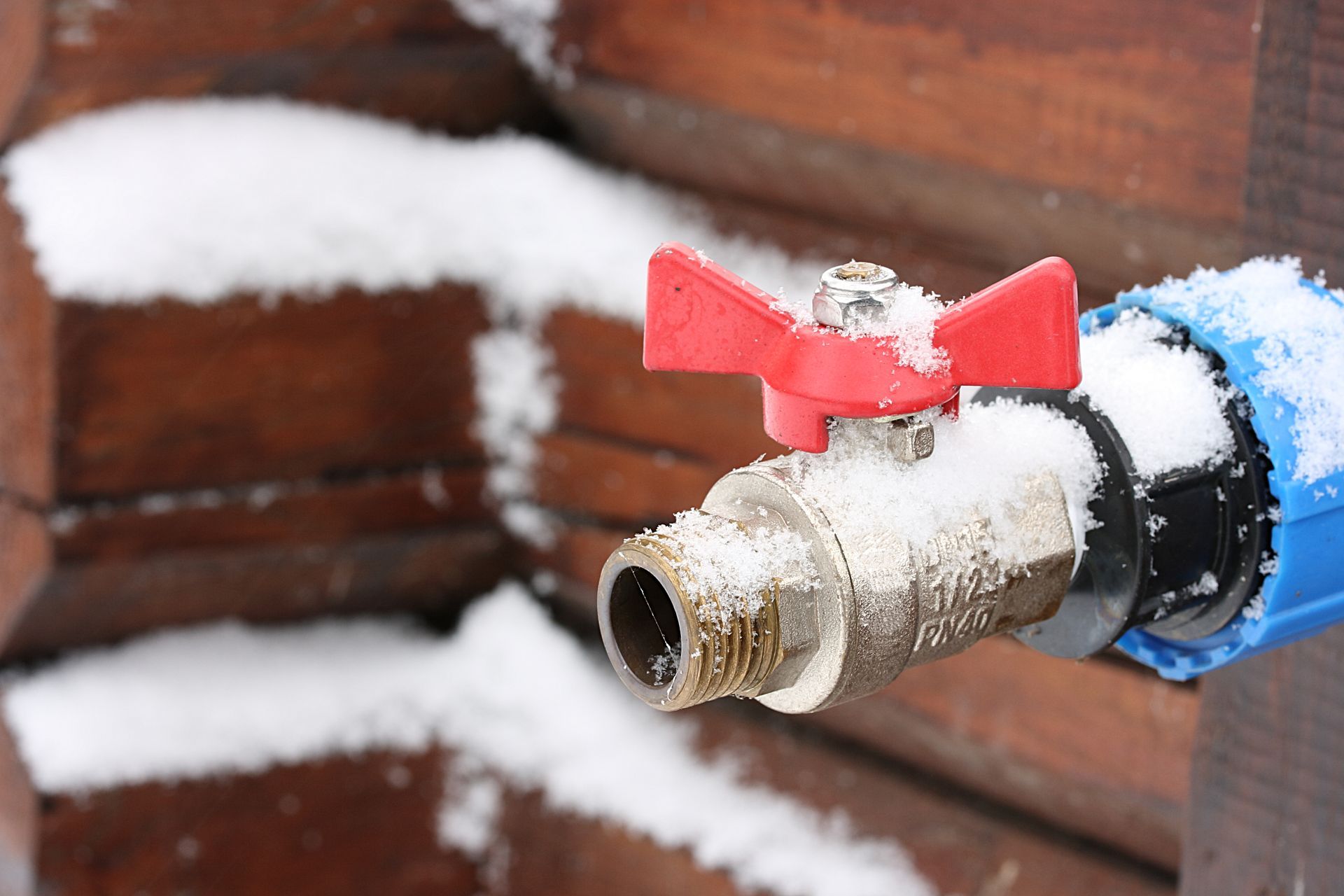 Snow-covered outdoor spigot with red handle on a wooden building.