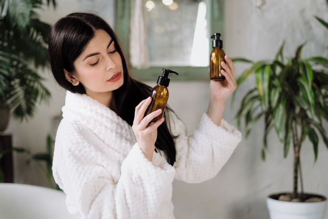 A woman in a bathrobe is holding two bottles of shampoo in her hands.