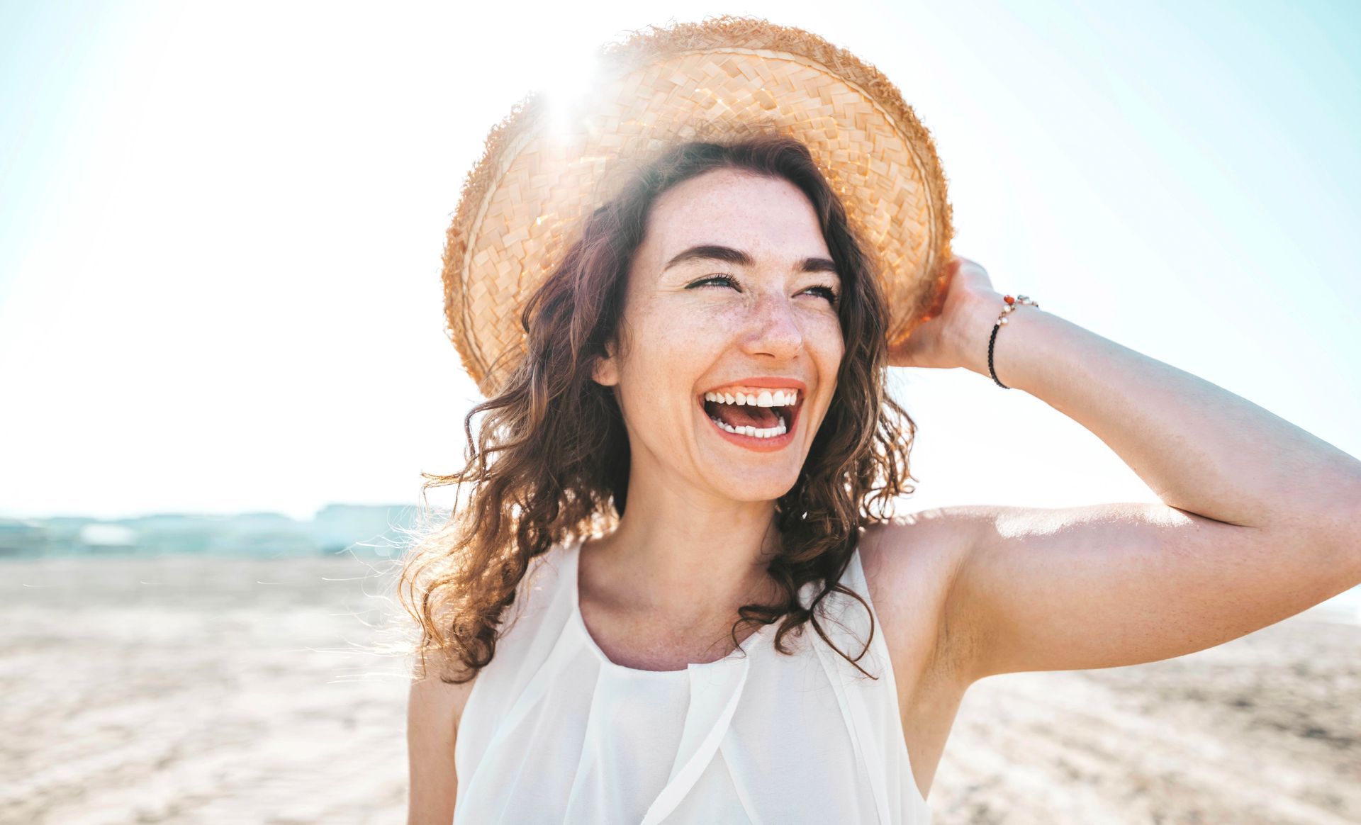 A woman wearing a straw hat is laughing on the beach.