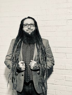 Professional headshot of Bradley McElrath, in a suit and tie, with long dreadlocks and tattoos, against a white brick wall.