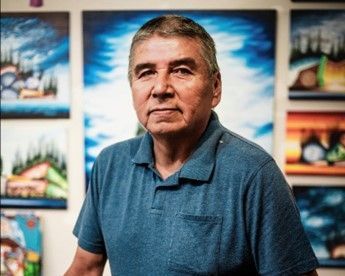 A man in a blue shirt is standing in front of a wall of paintings.