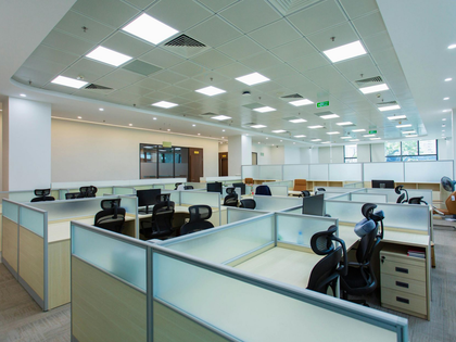 An open-plan office workspace featuring rows of cubicles with light wood desks, dark ergonomic chairs, and glass dividers.