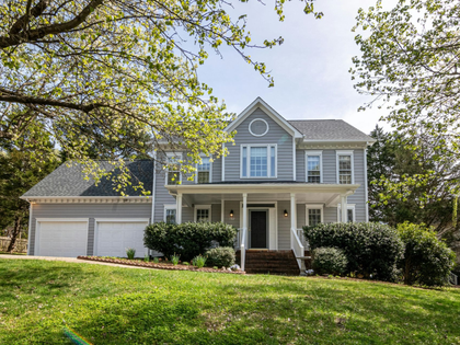 A light blue, two-story suburban house with a white front porch, a two-car garage, and green trees in a sunny front yard.