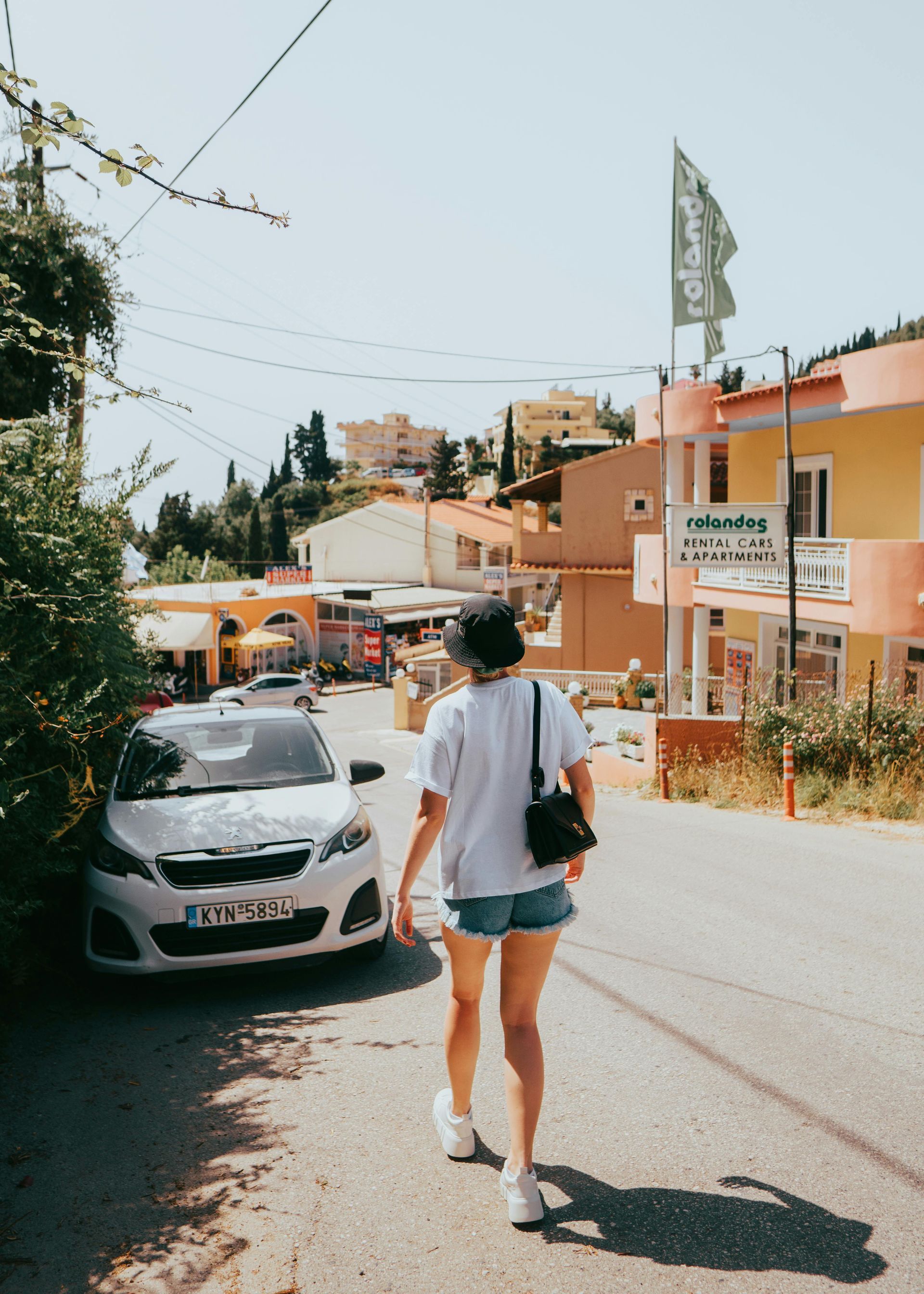 Person walks down a sunny street past parked car and colorful buildings.