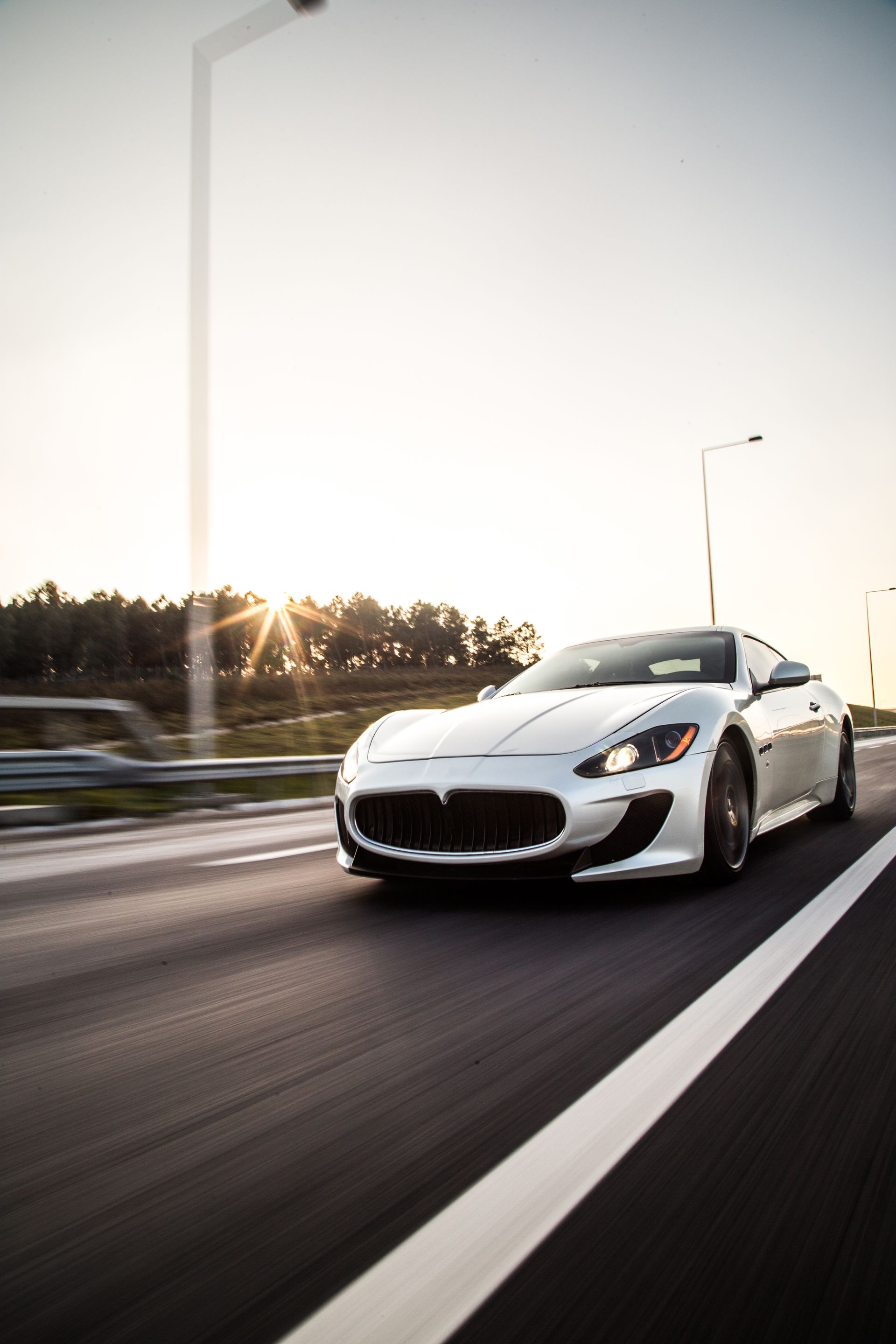 White Maserati sports car driving on a highway, sun in the background.