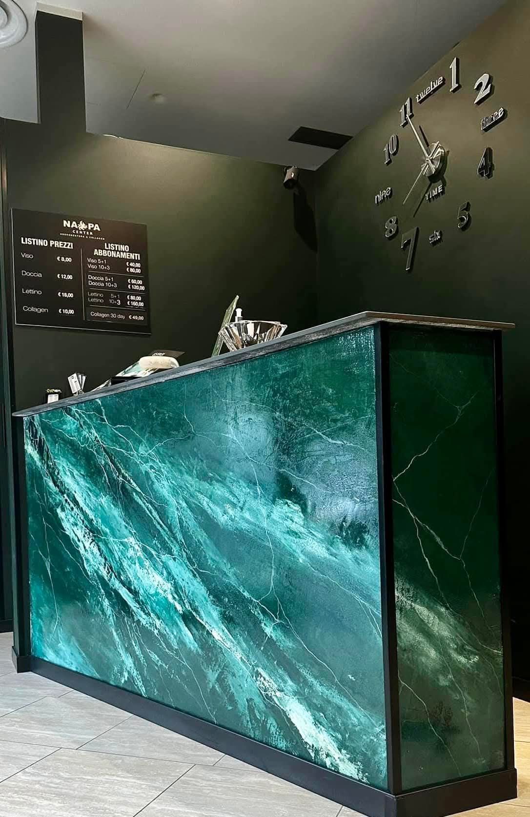 Banco reception con frontale in marmo verde in un interno moderno con pareti scure e pavimento piastrellato.