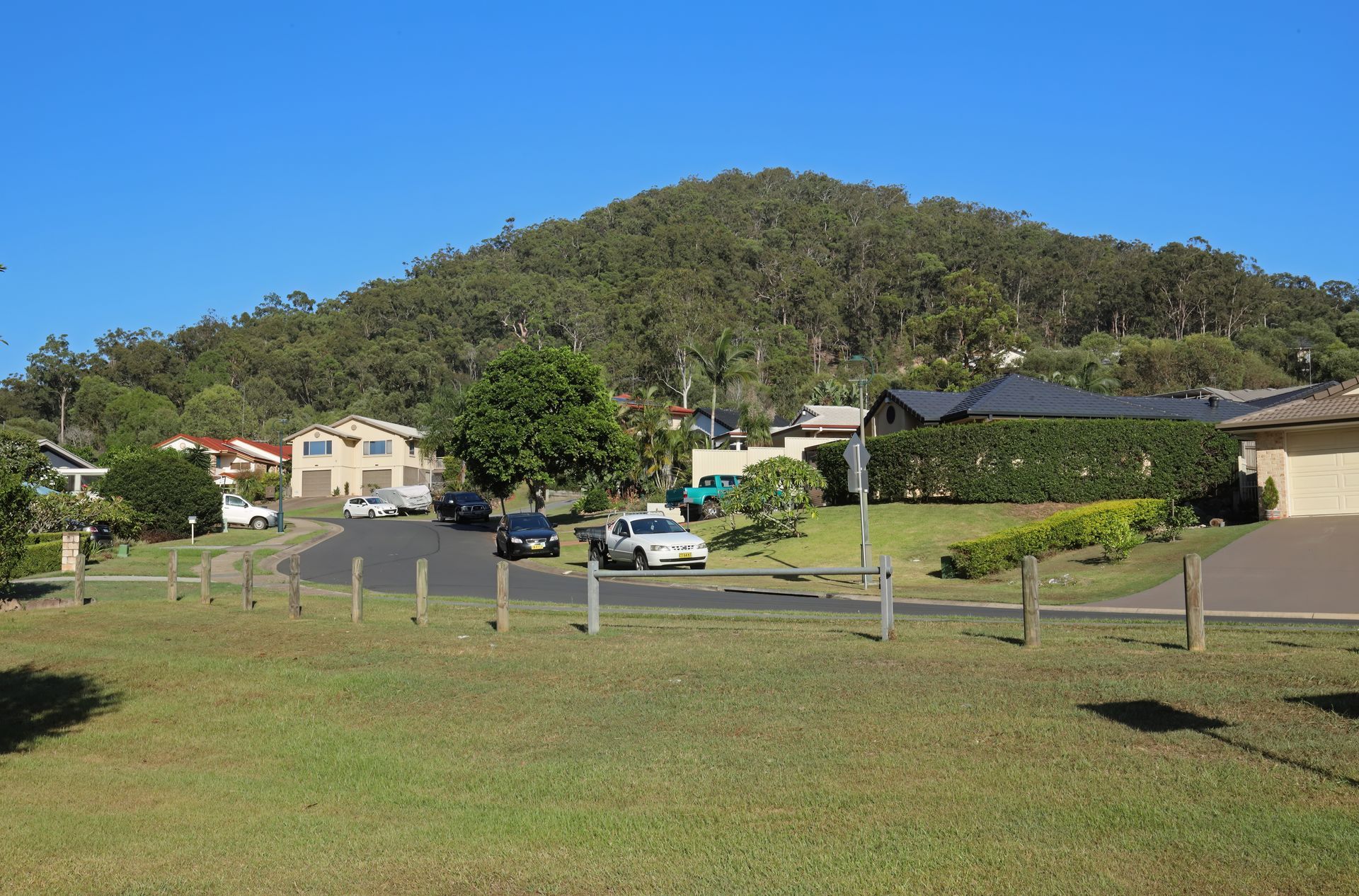 A grassy foreground leads to a suburban street with parked cars and houses, set against a large, tree-covered mountain — Leak N Pool In Logan, QLD