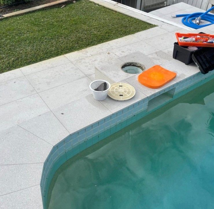 A pool skimmer lid is removed next to an open pool skimmer well on a paved deck with tools and a hose nearby.