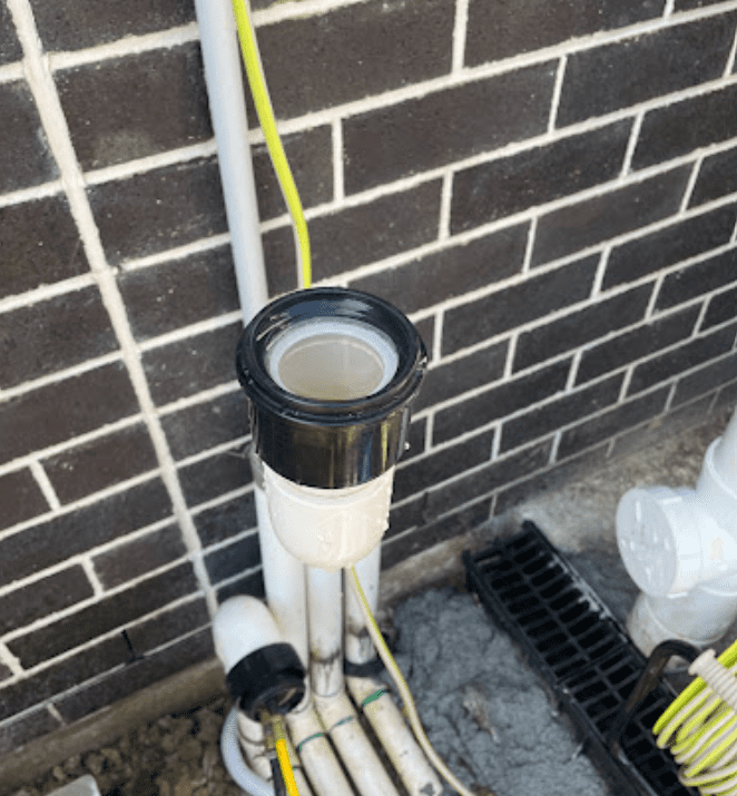 An open vertical plumbing pipe with a black rim and white base attached to a brick wall with other conduits nearby.