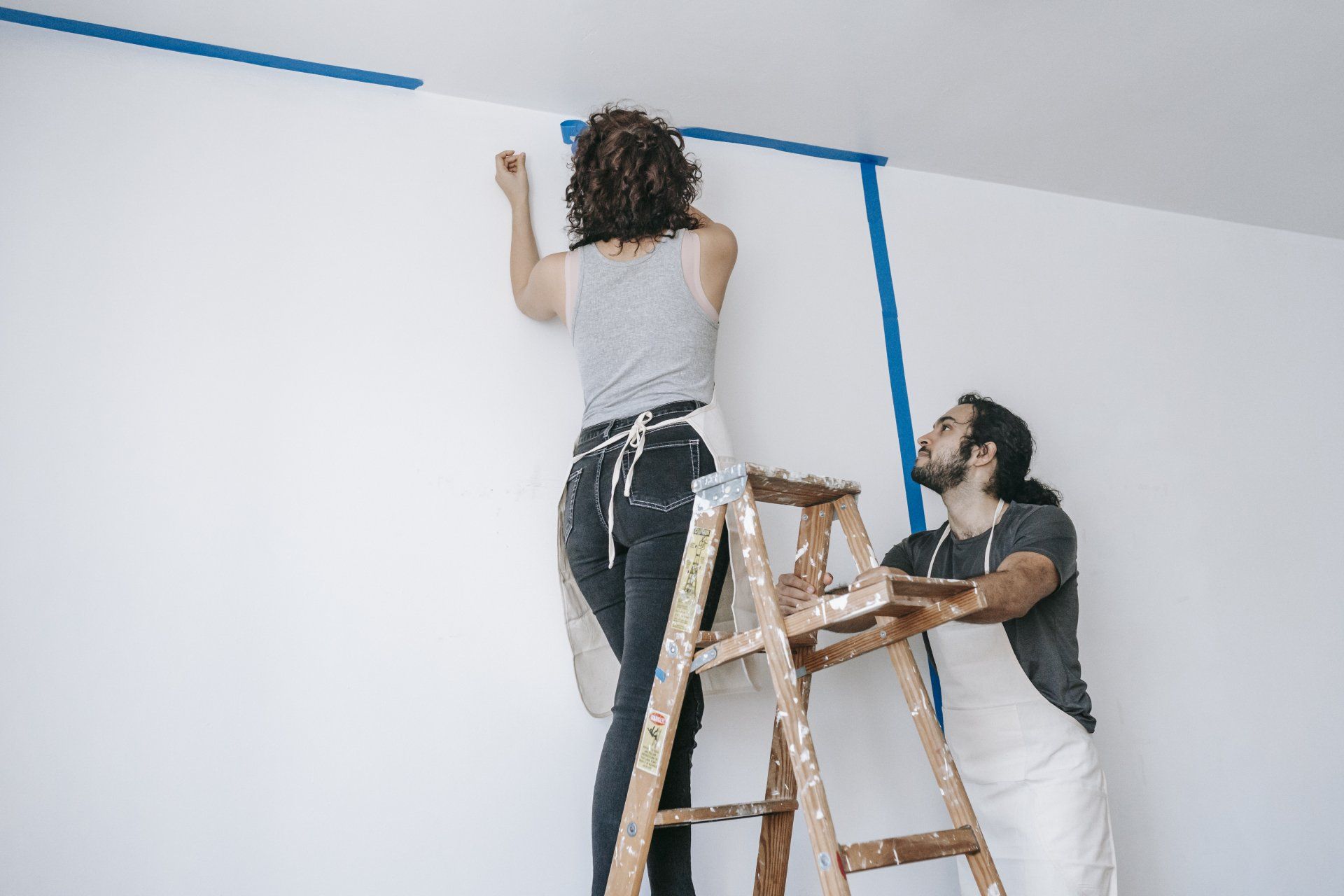 The picture is of two people putting up painter's tape on a ladder inside their home.