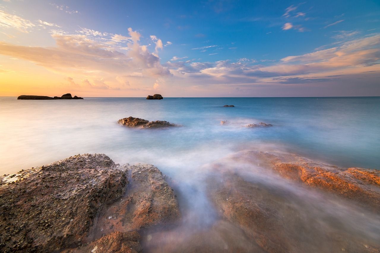 A serene image of the sky with clouds hovering over rocks that feed into the sea. 