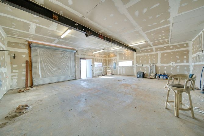 A spacious, unfinished garage with bare drywall, concrete flooring, a large overhead door, and a single wooden bar stool.