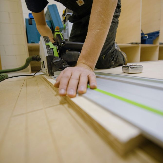 A person kneels on a workshop floor, using a green and black track saw to cut a piece of plywood.