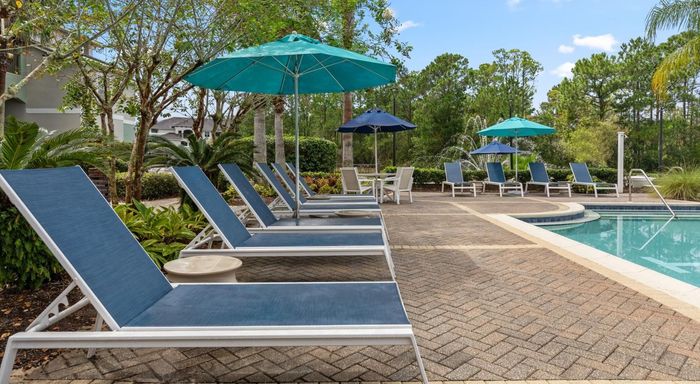 A swimming pool surrounded by a stone patio and lounge chairs, with palm trees and a residential building in the back.