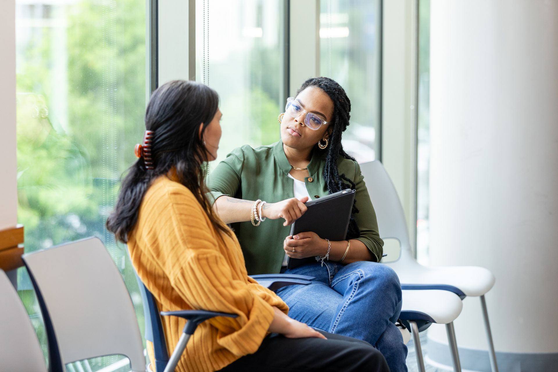 Two women are sitting in chairs talking to each other in front of a window.