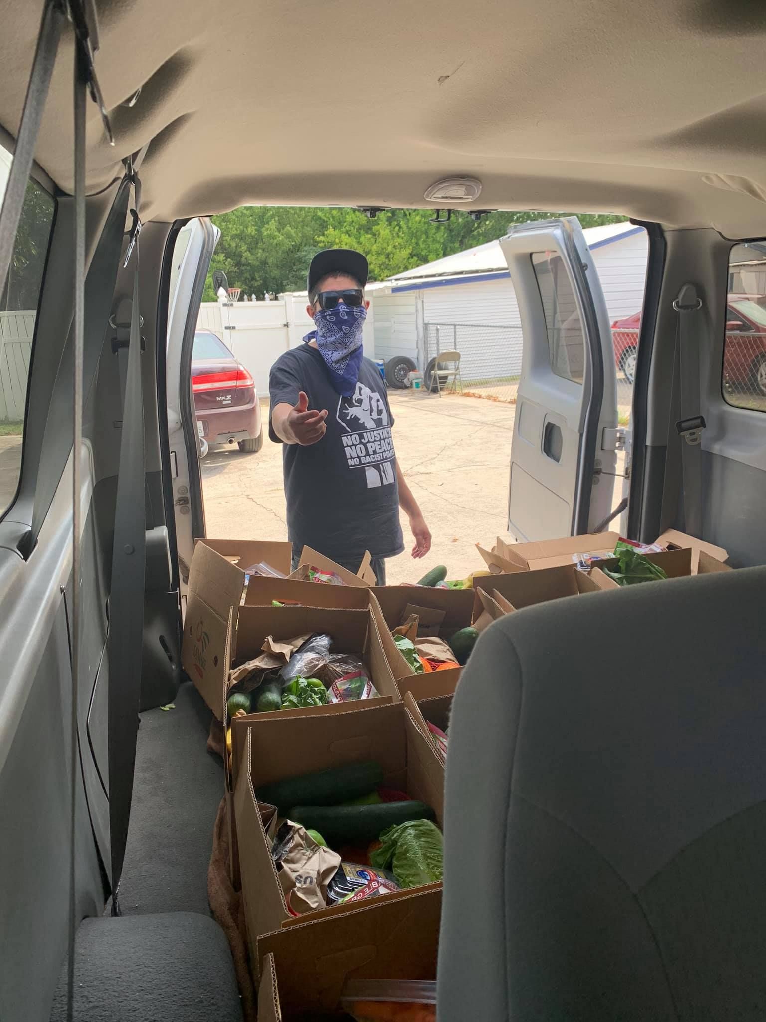 Person in van distributing produce boxes.  Boxes are filled with various vegetables. Outdoors on a sunny day.