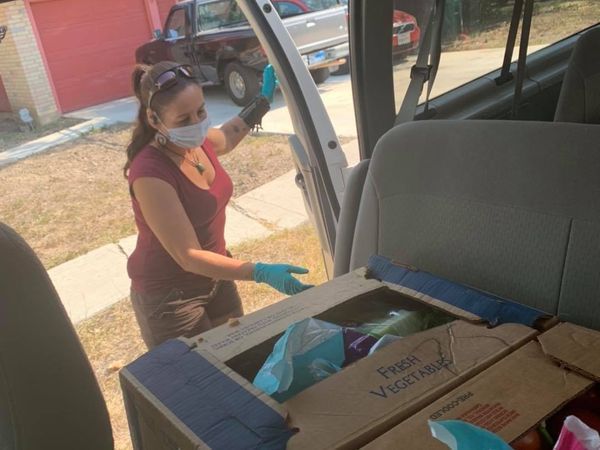 Woman in mask, gloves, loading vegetables into a van. Boxes, groceries. Outdoors, sunny.