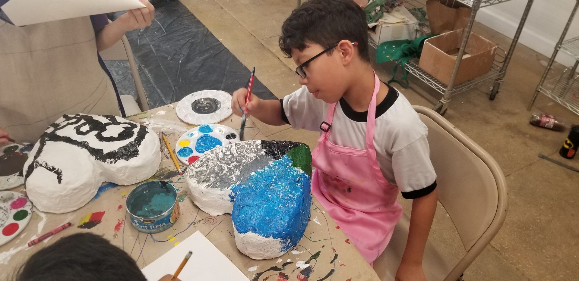 Boy with glasses and pink apron painting a mask at a table.