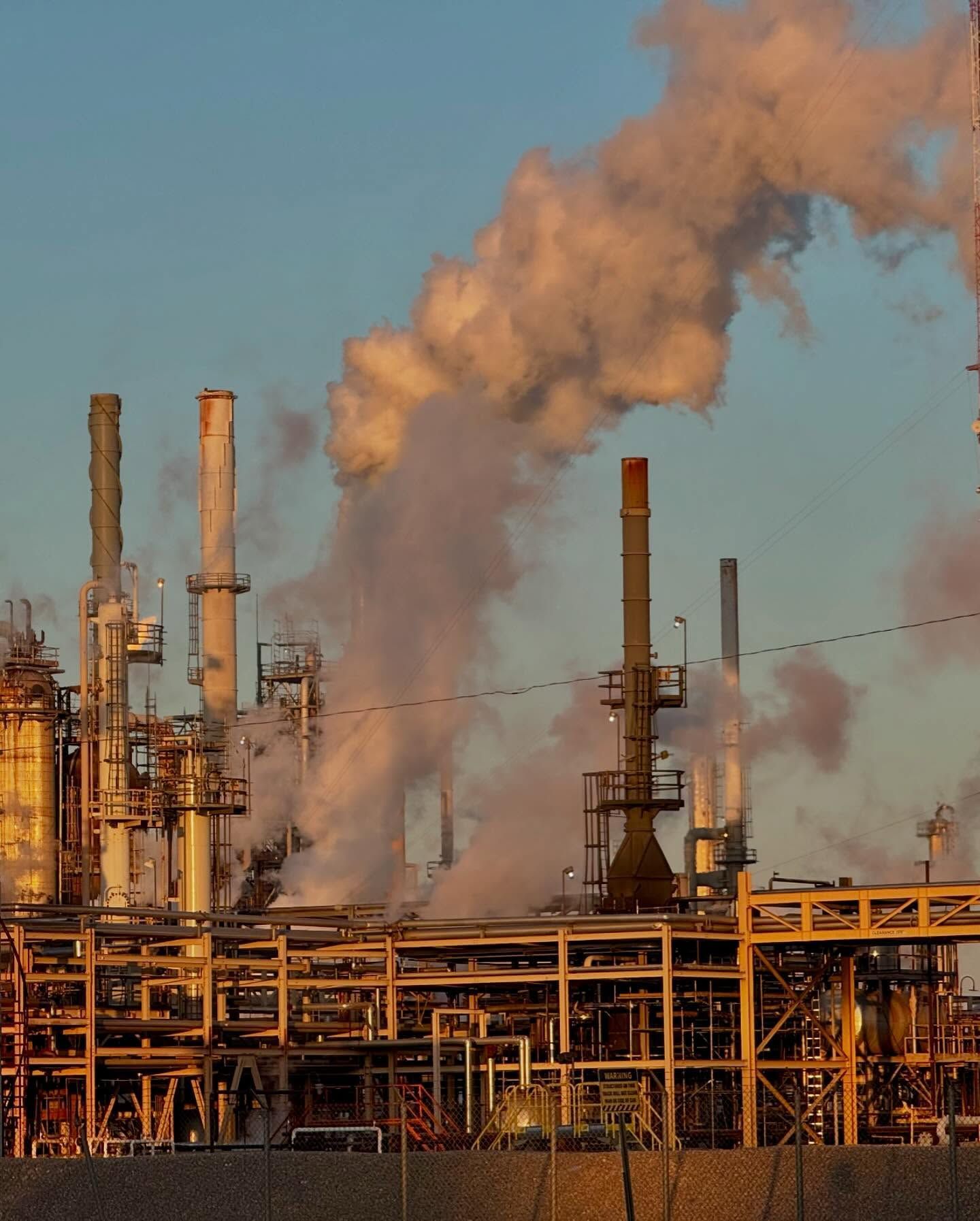 Oil refinery with smokestacks emitting white smoke against a blue sky; industrial setting.