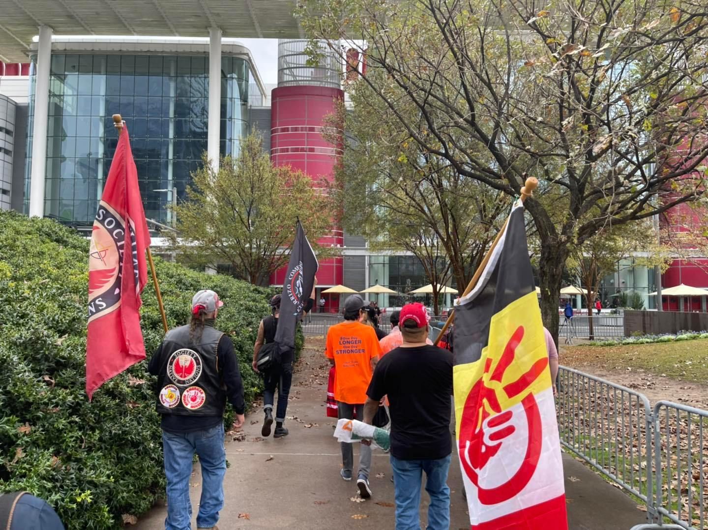 People march with flags outside a red and glass building, likely a protest or demonstration.
