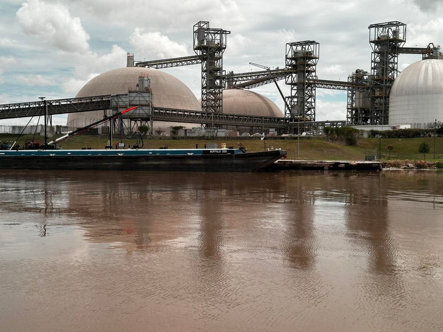 Barge on a muddy river near an industrial plant with dome-shaped storage tanks and conveyor structures.