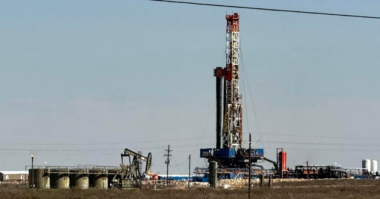 Oil drilling rig in a field, with tanks and pumping units, under a blue sky.