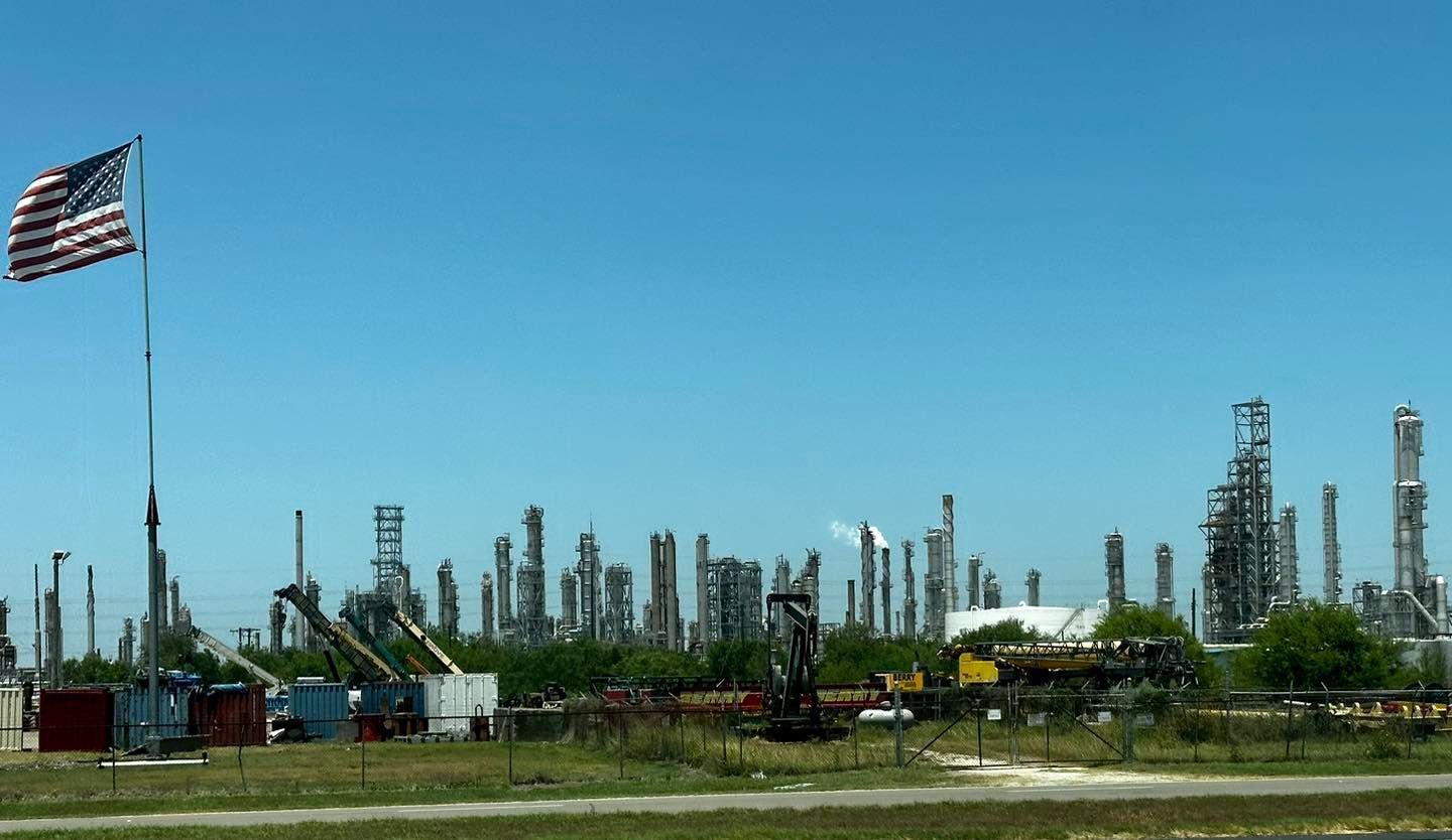 American flag waving over an oil refinery under a blue sky.