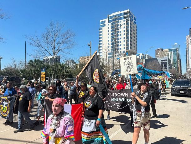 Indigenous people march in a city, holding signs and flags, protesting. Bright sunny day.