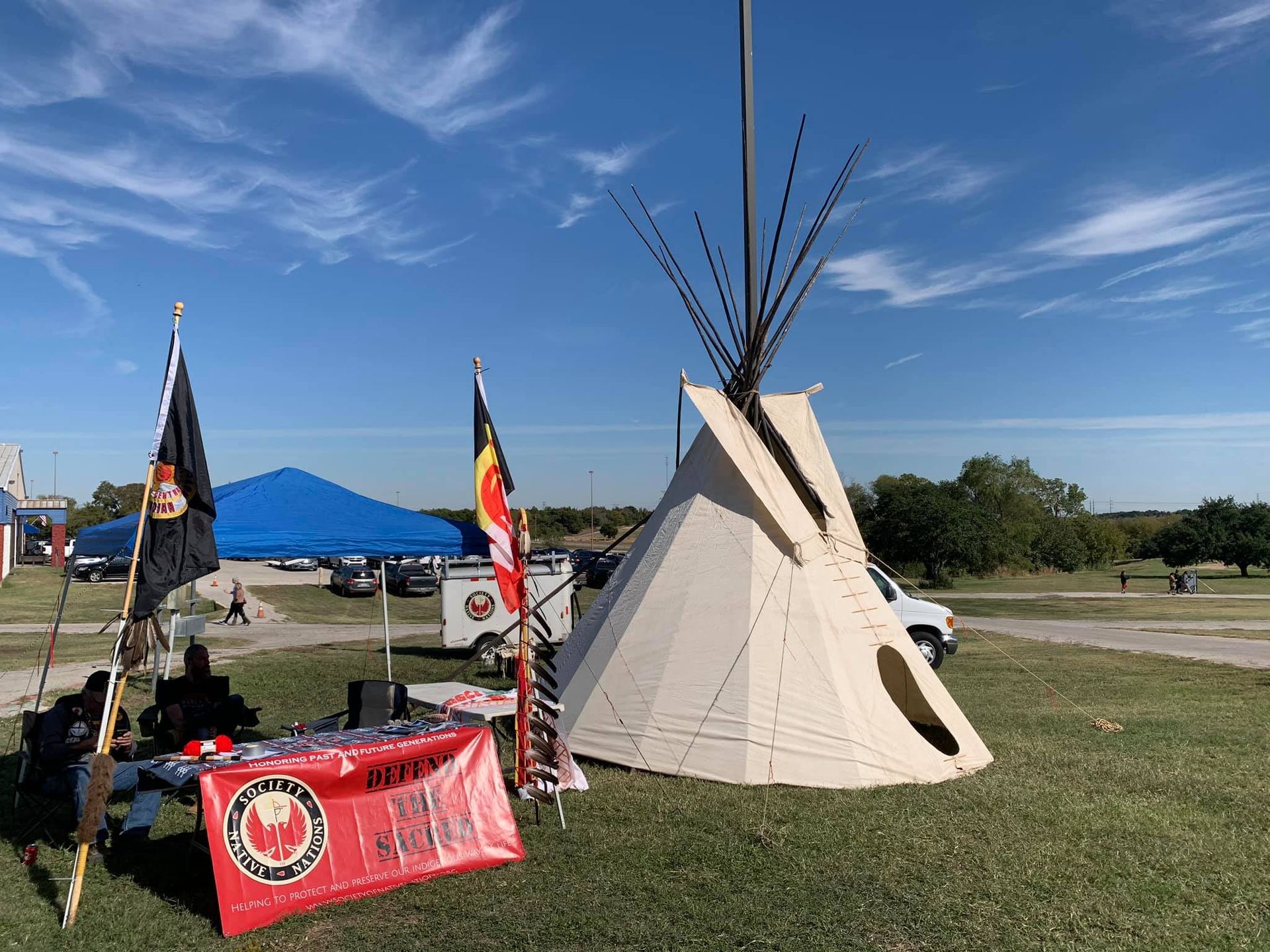 A tipi set up at an outdoor event with flags, a table, and a blue tent on a grassy field.