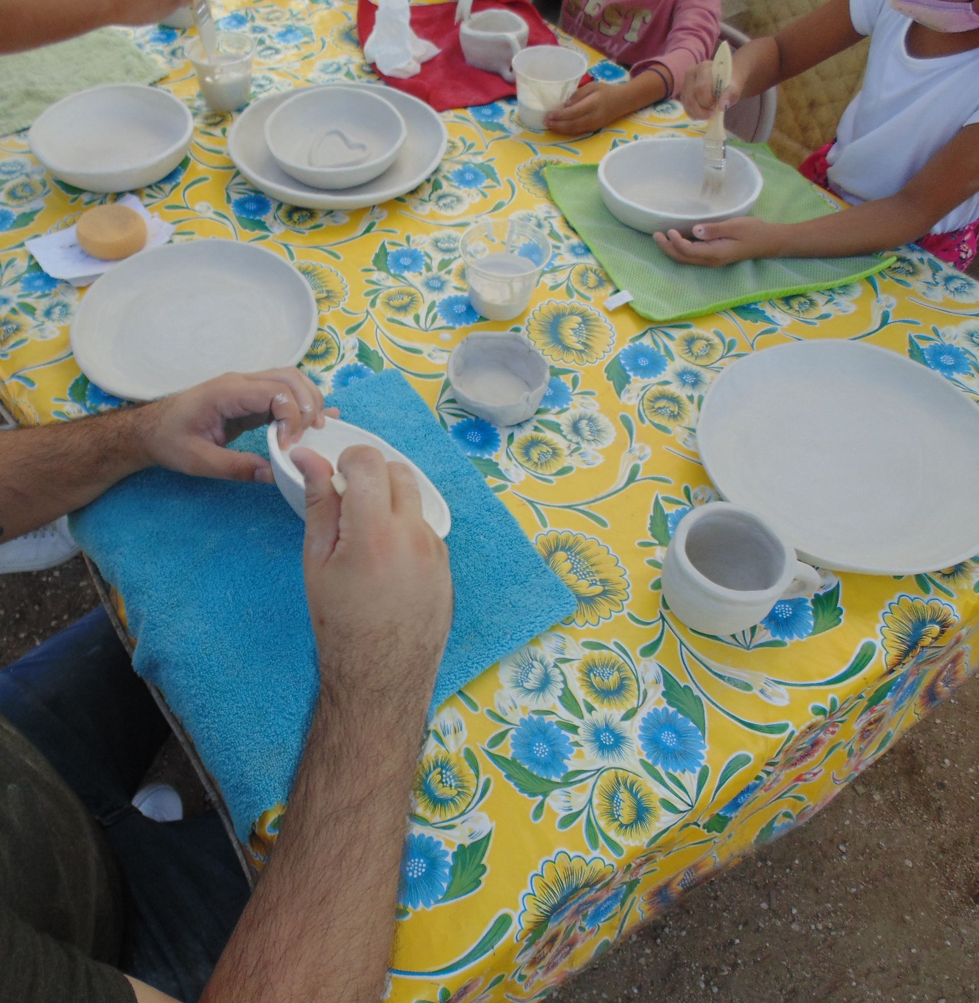 People at a table doing pottery. Yellow floral tablecloth, hands holding bowls.