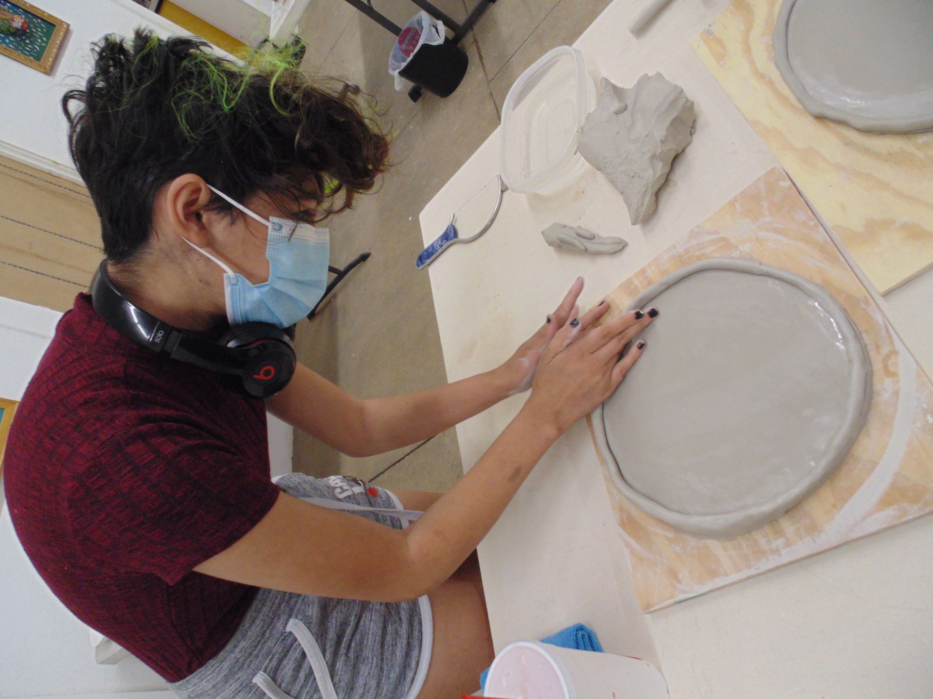 Person wearing a mask molding clay into a circle. Table with other clay pieces and tools.