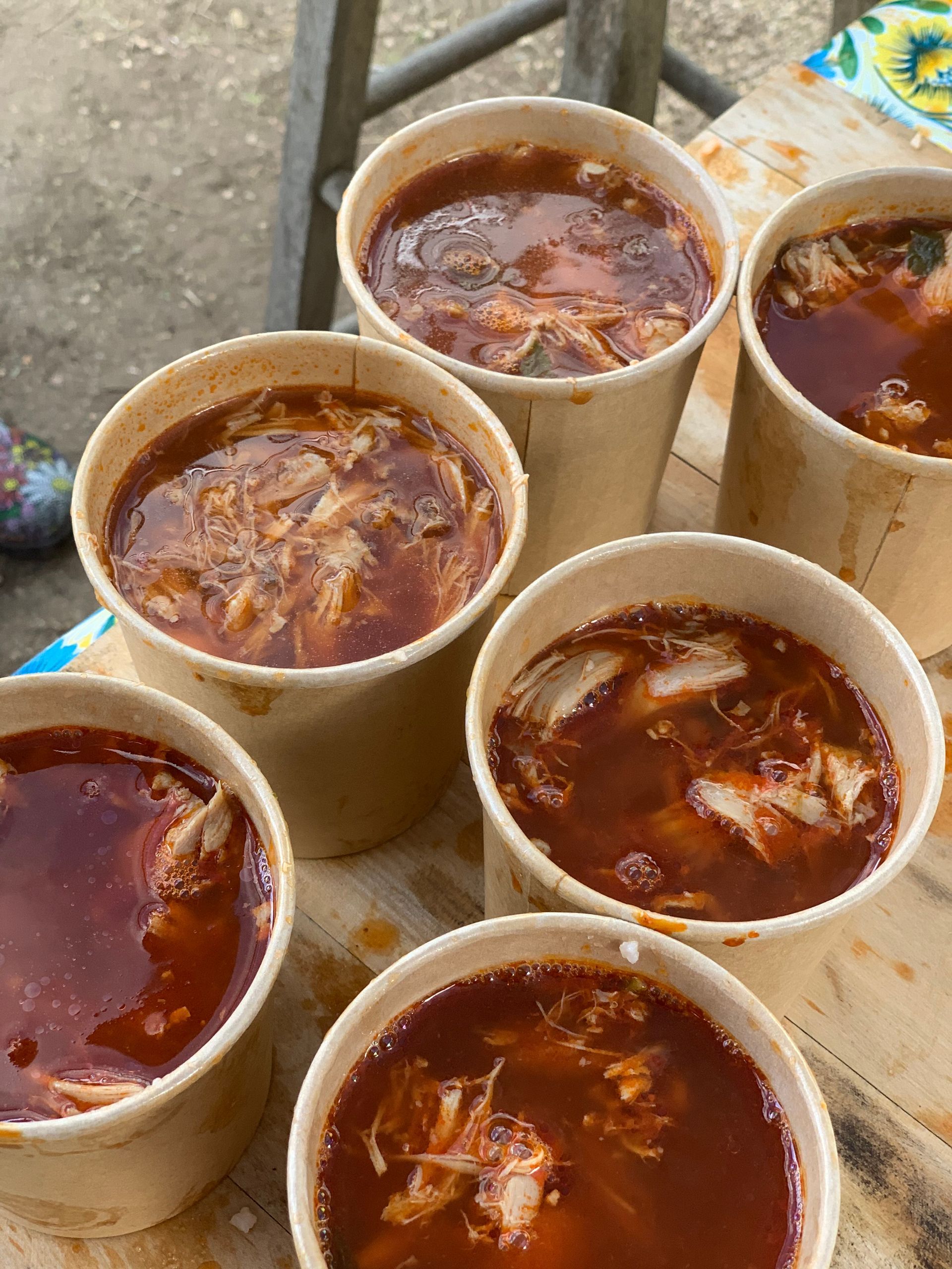 Six paper cups filled with a reddish-brown liquid, possibly soup or stew, outdoors.