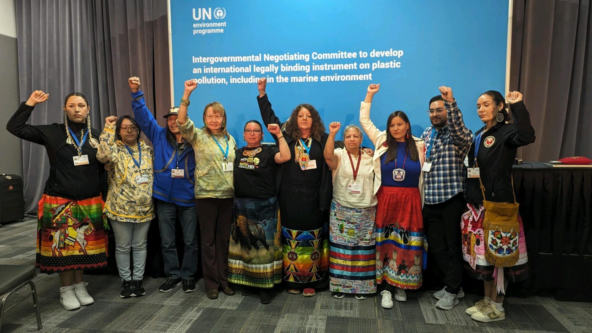 Indigenous people raising fists in front of a UN banner; indoors.