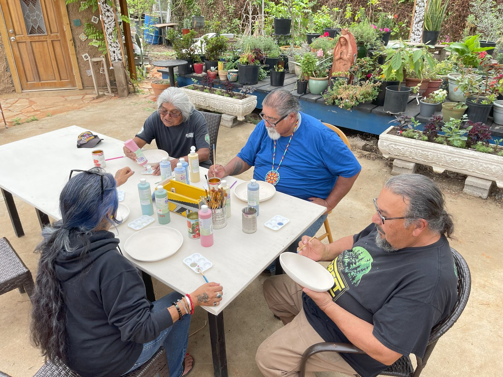 Four people painting ceramic plates at a table outdoors, surrounded by plants.