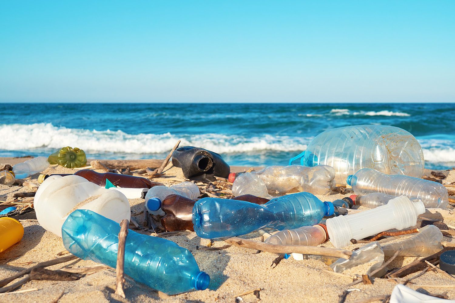 Plastic bottles and trash litter a sandy beach, waves in the background.