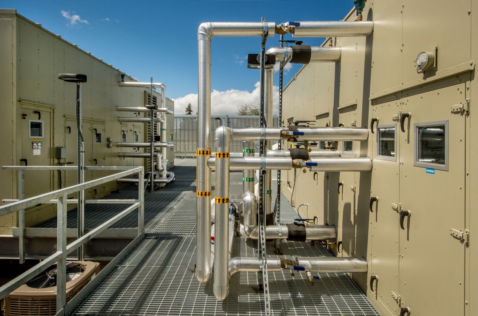Rooftop mechanical equipment with metallic pipes, walkways, and blue sky.