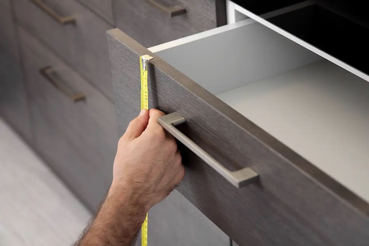 Person measuring an open gray drawer with a yellow tape measure in a kitchen.
