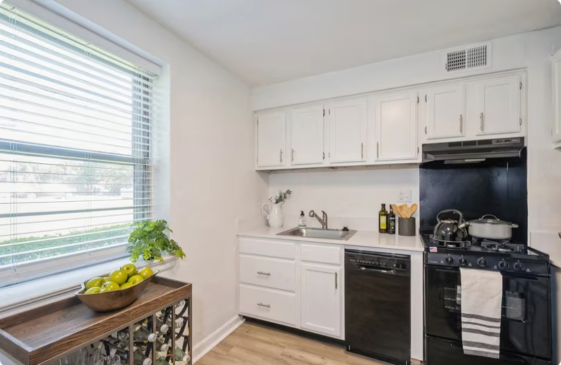 Small white kitchen with black appliances, a window with blinds, and a wooden table.